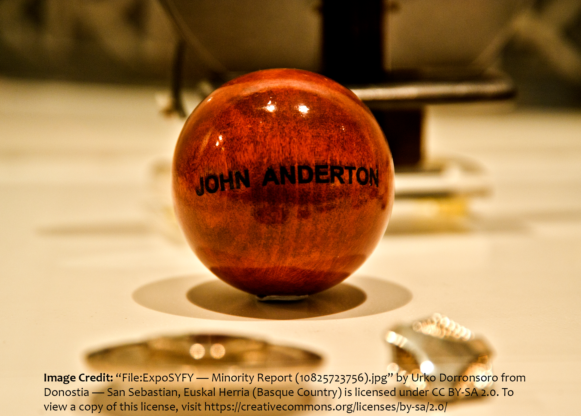 Closeup photo of a small, dark-stained, polished wooden ball with the name, 'John Anderton,' carved into it and darkened with black paint. From the movie, 'Minority Report.' Credited to Urko Dorronsoro on Creative Commons.