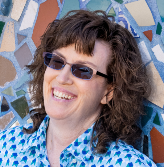 Headshot shows a White woman with an open smile, wearing dark, blue-rimmed glasses. Her hair is medium length and wavy-brown. Her shirt shows various shades of blue in a geometric chevron pattern. Her head appears to be leaning backwards against a wall mosaic made of colored tile shards against an aqua background.